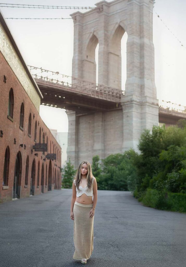 A high school senior with long blonde hair stands on a quiet street beneath the Brooklyn Bridge, flanked by a red brick building and green foliage. She is wearing a white crochet top and beige crochet skirt, walking towards the camera with a calm expression.