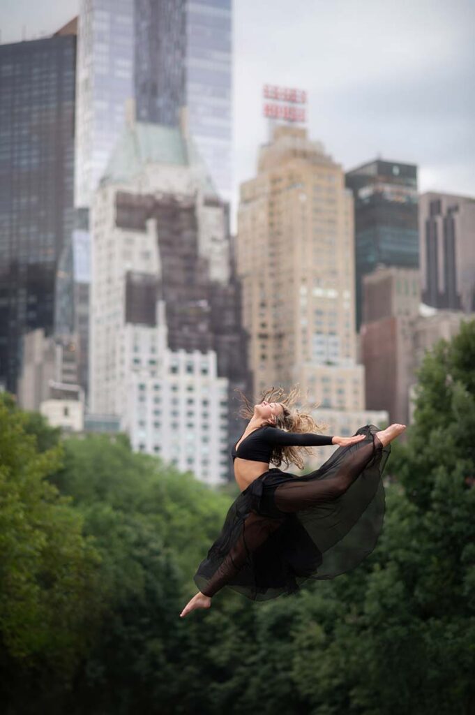 High school senior dressed in a black outfit gracefully leaps mid-air in a park, surrounded by lush green trees and towering city buildings. The dynamic movement captures the essence of an NYC senior photoshoot, as the urban skyline contrasts beautifully with the natural setting.