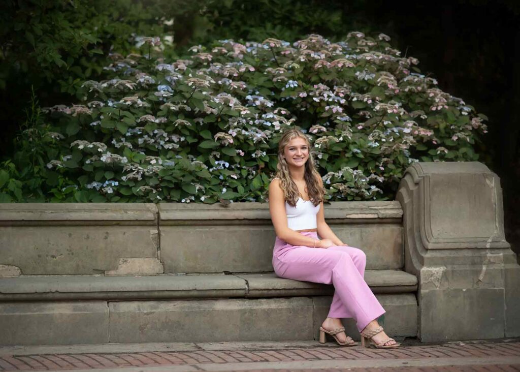 A teen with long hair, wearing a white crop top and pink pants, is sitting on a stone bench in front of a large hydrangea bush. She is smiling and appears relaxed in the outdoor setting, capturing the essence of a serene senior portrait in NYC.
