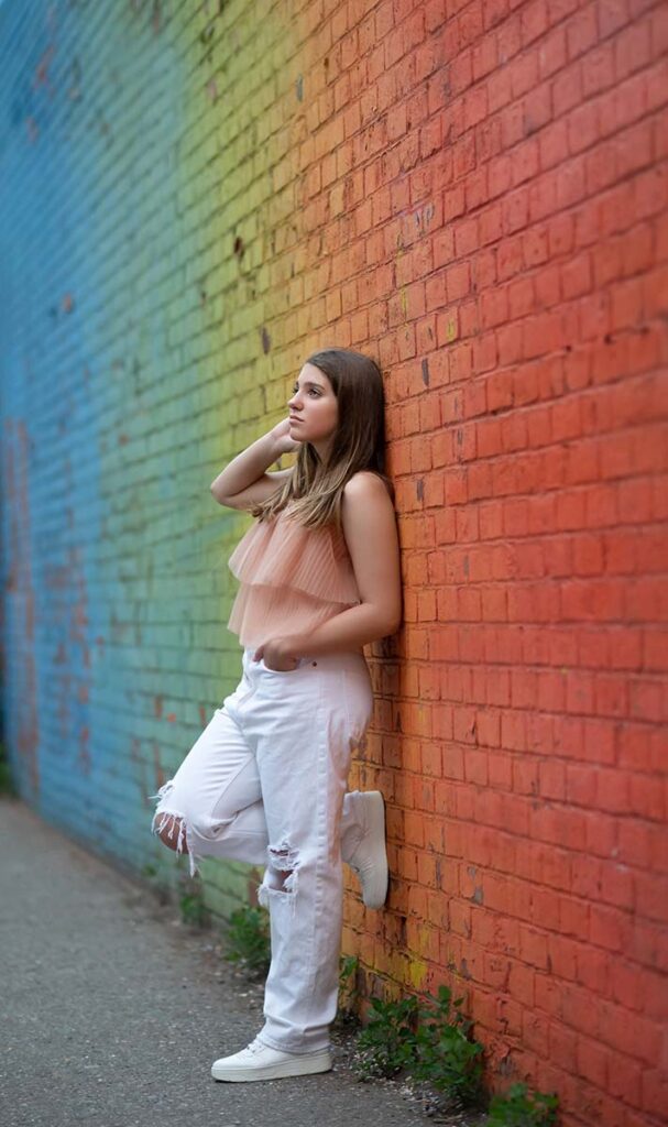 A high school senior with long brown hair leans against a colorful brick wall, with hues transitioning from blue to green to red. She is wearing a light pink sleeveless top and white ripped jeans, and one leg is bent with her foot against the wall.
