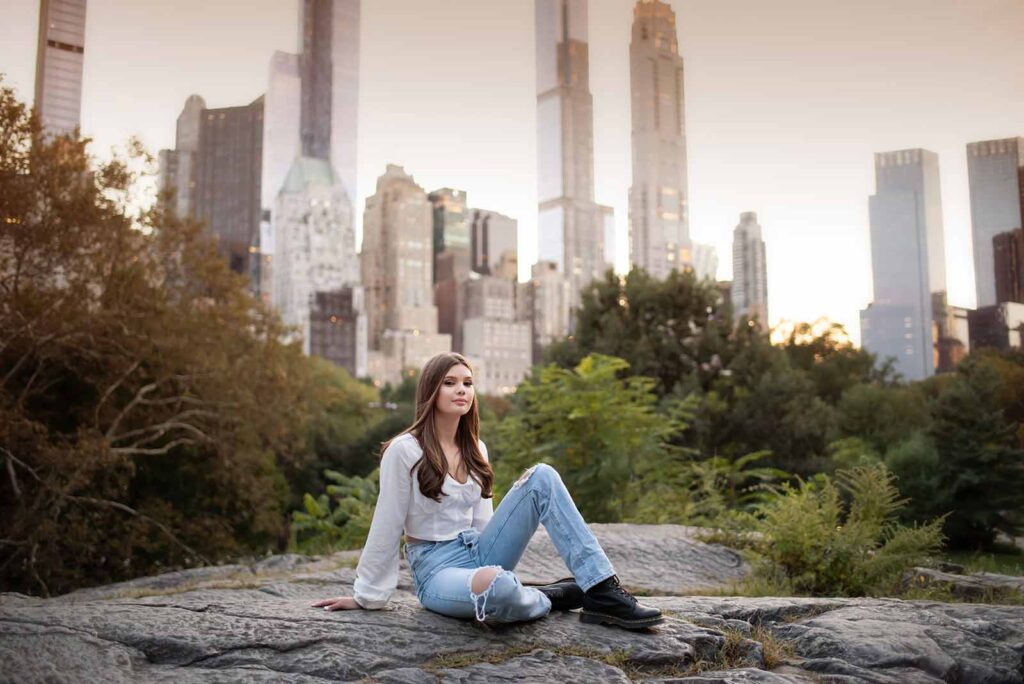 A senior in a white shirt and ripped jeans sits on a large rock in a park, with the NYC skyline featuring tall modern buildings in the background. She has long hair and is looking slightly to her right, surrounded by greenery under a serene sky.