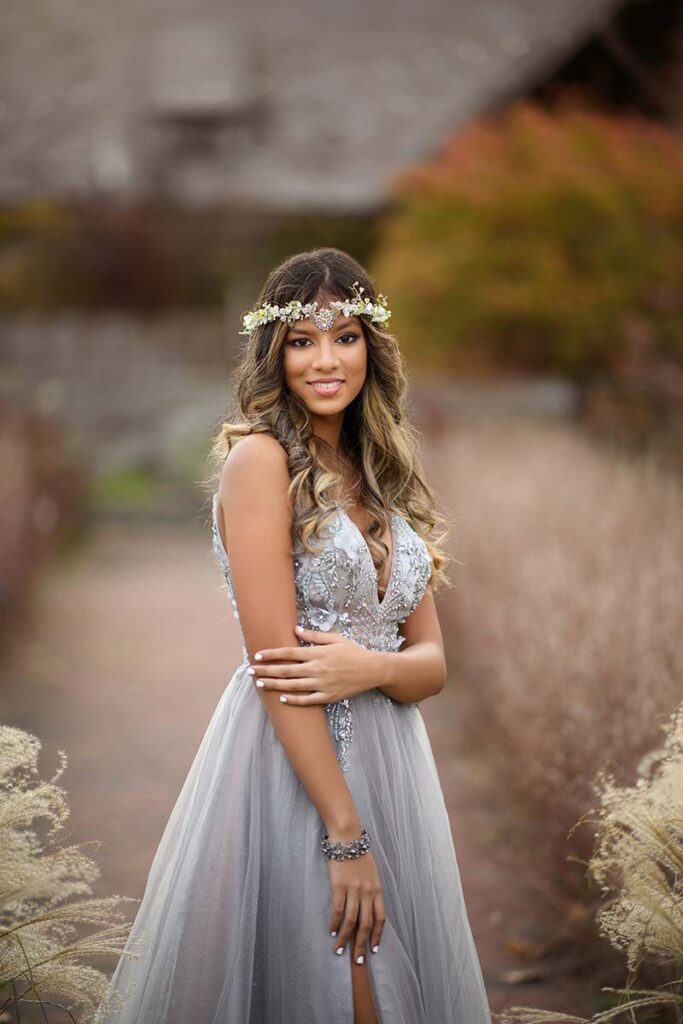 A teen poses outdoors for a stunning senior portrait, wearing a long, light gray, sleeveless dress adorned with sequins and a thigh-high slit. She has long, curly hair and wears a floral headband. Smiling brightly, she stands amidst tall grasses with blurred foliage in the background.