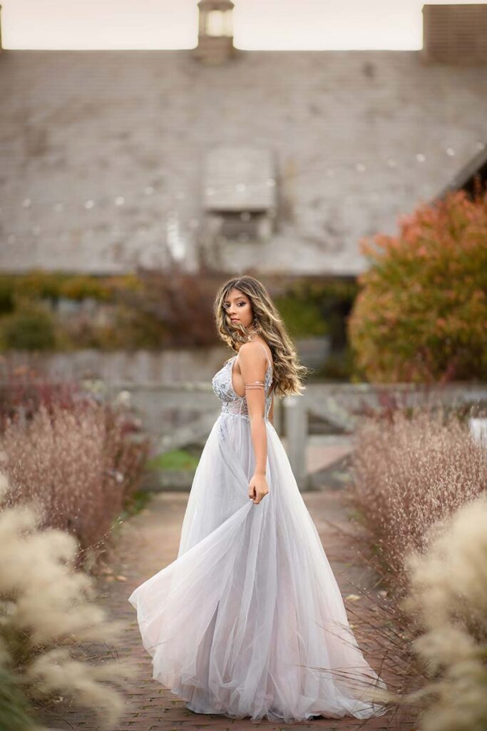 A teen with long hair stands outdoors on a brick path lined with tall grasses. She wears a flowing, light-colored gown and looks over her shoulder towards the camera, capturing the perfect moment for a high school senior photoshoot. The background features a building with a large sloped roof and autumnal foliage.