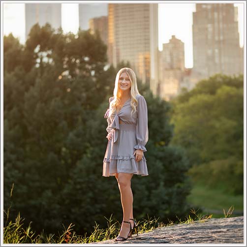 A blonde teen in a light gray dress and black heels stands on a grassy area in Central Park with tall trees and city buildings in the background. The sun is setting, casting a warm glow over her senior portrait scene.