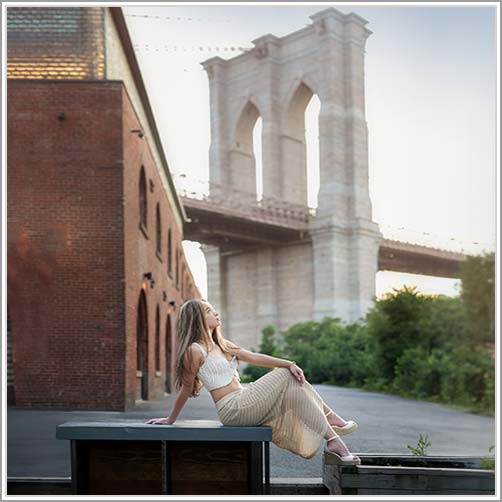 A high school senior with long hair wearing a white crop top and beige skirt sits on a ledge, looking up and smiling. Behind her is a red brick building and the Brooklyn Bridge, captured during daylight. The greenery checks out beautifully against the urban landscape.
