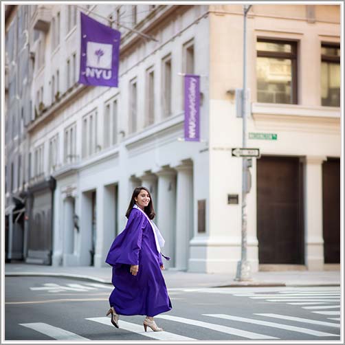 NYU graduate in a purple graduation gown and cap walks across a crosswalk in an urban setting. Two vertical banners with the NYU logo are visible on the buildings in the background. The joyful graduate, likely posing for their NYU senior portrait, smiles while looking back.