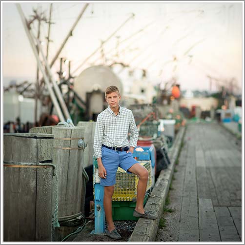 A young man stands on a dock at South Street Seaport, leaning against a lobster trap. He is wearing a checkered shirt, blue shorts, and boat shoes. The background features fishing equipment and boats, with a blurred effect creating a rustic and serene atmosphere.