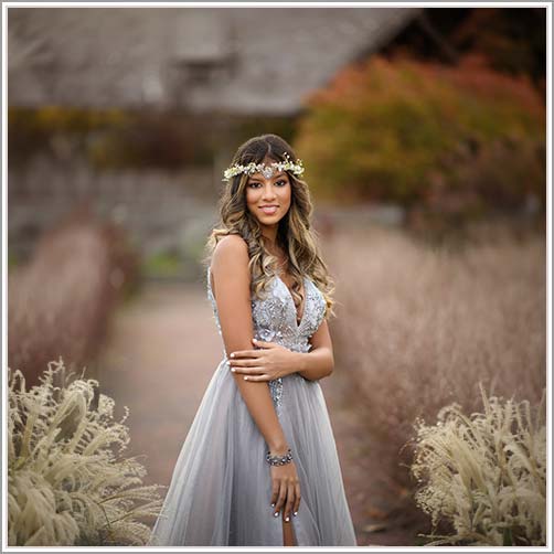 A young lady wearing a silver gown adorned with floral embellishments and a matching floral headpiece stands in a lush garden. With long, wavy hair and a soft smile, she poses gracefully for her Westchester County senior portrait against a backdrop blurred with greenery and autumnal hues.