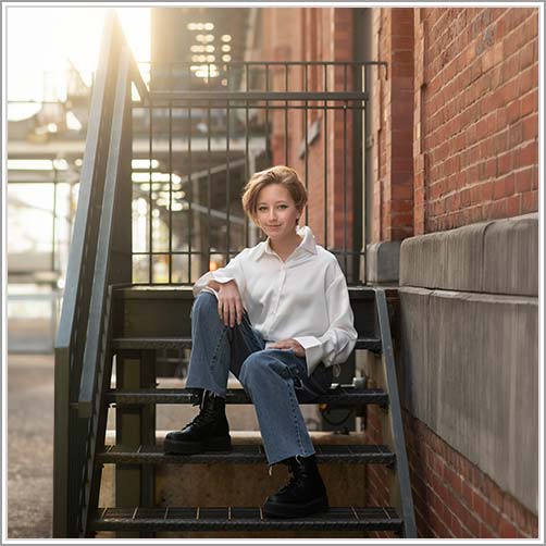 High school senior with short hair sits on an outdoor staircase in Tribeca, NYC, wearing a white shirt, blue jeans, and black boots. The background features an industrial setting with brick walls and metal structures. The sunlight creates a warm glow—perfect for a senior photoshoot.