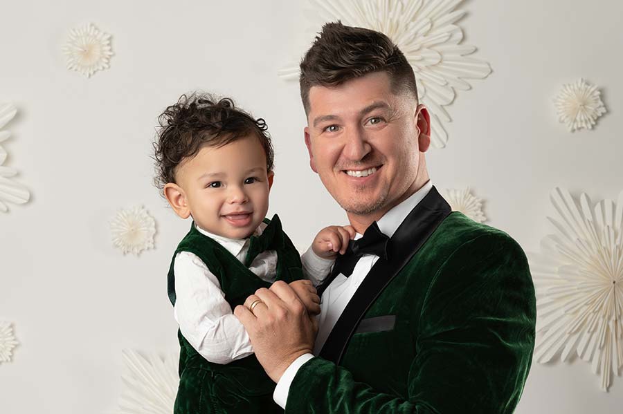 A man with short dark hair, wearing a green velvet suit and bow tie, smiles while holding a young child in a matching green velvet outfit. Both are looking at the camera in a NYC photo studio backdrop decorated with holiday theme.