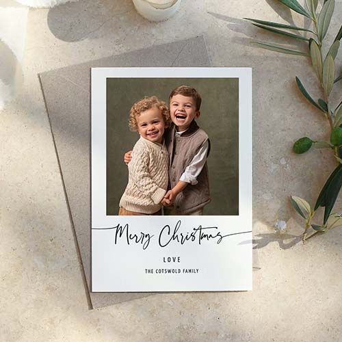 A holiday card featuring two smiling children, one with curly blond hair in a knit sweater and the other with straight brown hair in a vest. The card says 