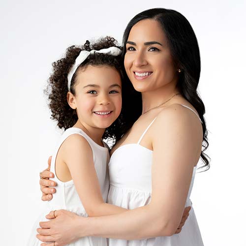 A woman and a young girl wearing matching white dresses smile while embracing. The woman has long dark hair, and the girl has curly hair with a white headband. Both are looking directly at the camera against a plain white background, capturing a heartwarming moment perfect for a Christmas photoshoot.