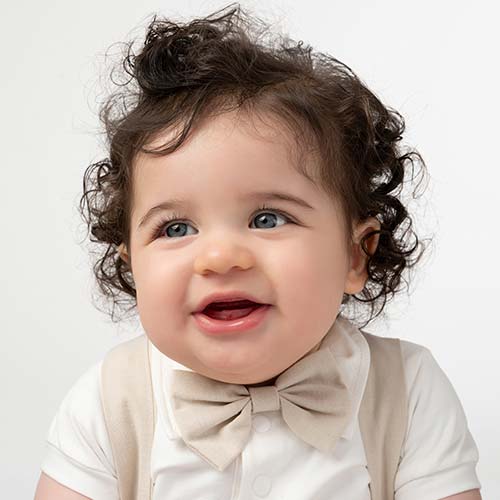 A smiling baby with curly hair wears a beige bow tie and matching beige vest over a white shirt. Captured by a top baby photographer in NYC, the simplicity of the plain white background highlights the adorable details.