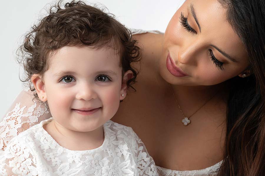 A woman with long dark hair and a flower necklace looks down lovingly at a smiling young child with curly hair. Both are wearing white lace outfits, capturing the essence of a heartfelt holiday photoshoot against a plain white background.