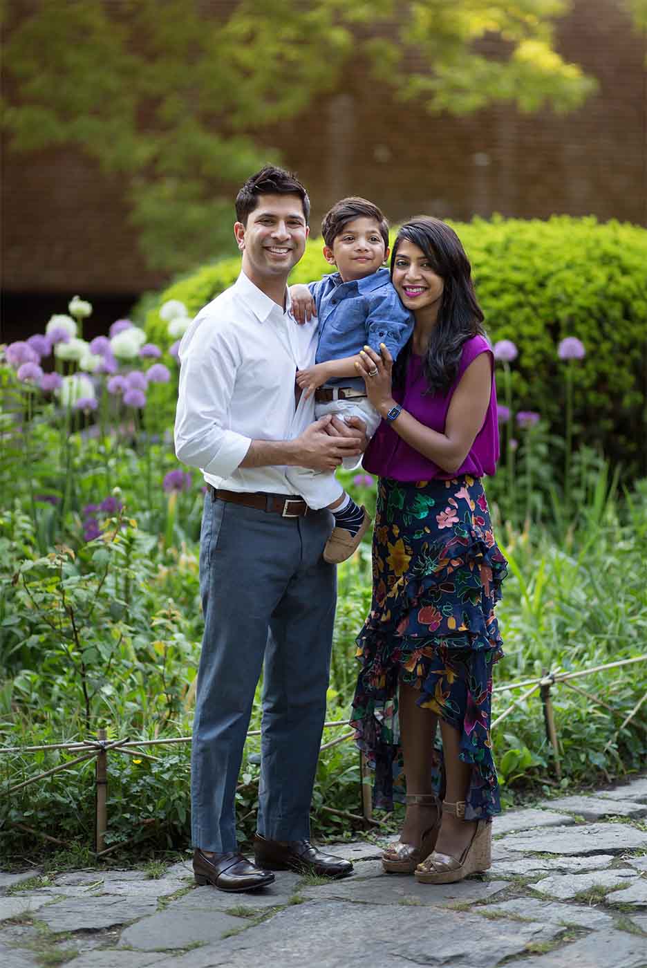 A husband and wife stand outdoors in Central Park with their young son. The father, dressed in a white shirt and gray pants, holds his son, who is wearing a blue shirt and gray pants. The mother, in a purple top and colorful skirt, stands beside them. They are surrounded by the lush greenery and flowers of the park.
