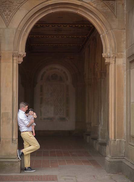 A father leans against a column under a stone archway in Central Park, holding his daughter who is looking outward. The archway frames them, leading into a dimly lit passage with detailed stonework and a patterned window at the end. The man and baby are casually dressed.