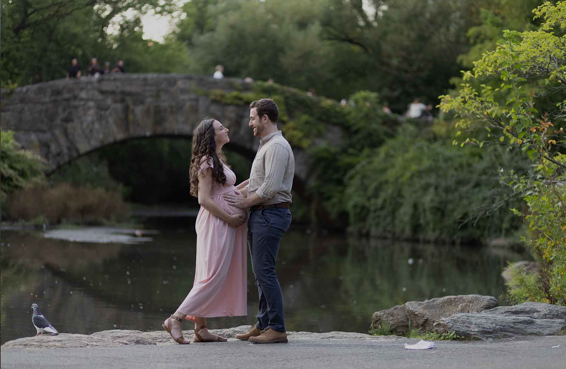 A pregnant woman in a flowing pink dress and a man in a light shirt and dark pants smile and hold hands, gazing at each other beside a calm pond. In the background, Central Park's stone bridge, lush greenery, and a pigeon on the path add to the serene ambiance.