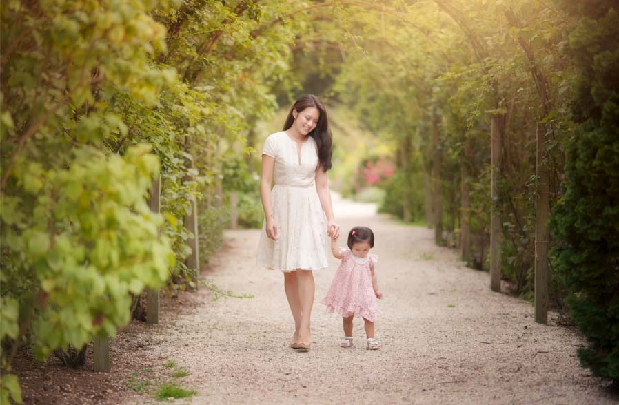 A woman and a young child in dresses walk hand in hand down a leafy garden path lined with green foliage. Sunlight filters through the arches overhead, creating a warm, serene atmosphere.