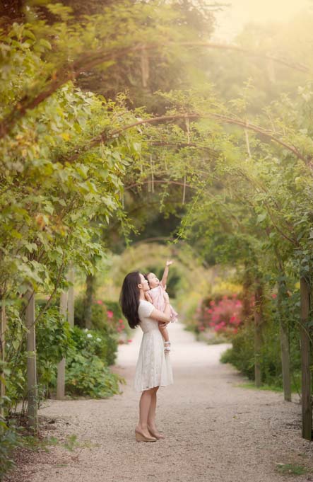 A woman in a white dress holds a child on a garden pathway, surrounded by lush greenery and vibrant flowers. It's the perfect setting for a heartfelt photoshoot, as they enjoy the serene atmosphere under the canopy of arching branches.