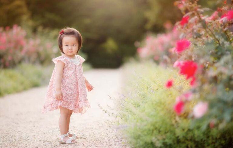 A young girl in a pink floral dress poses on a gravel path surrounded by vibrant, blooming flowers. The sun casts a warm glow on the scene, as if captured through the lens of a masterful photographer, highlighting the lush greenery and colorful blossoms.