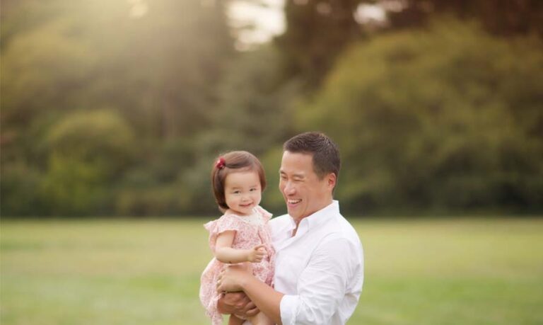 In a sunlit, grassy field, a father holds his smiling toddler daughter in his arms. Both are dressed in light, summery clothes and radiate joy. The scene captures a beautiful moment of photography with soft-focus greenery and trees in the background.