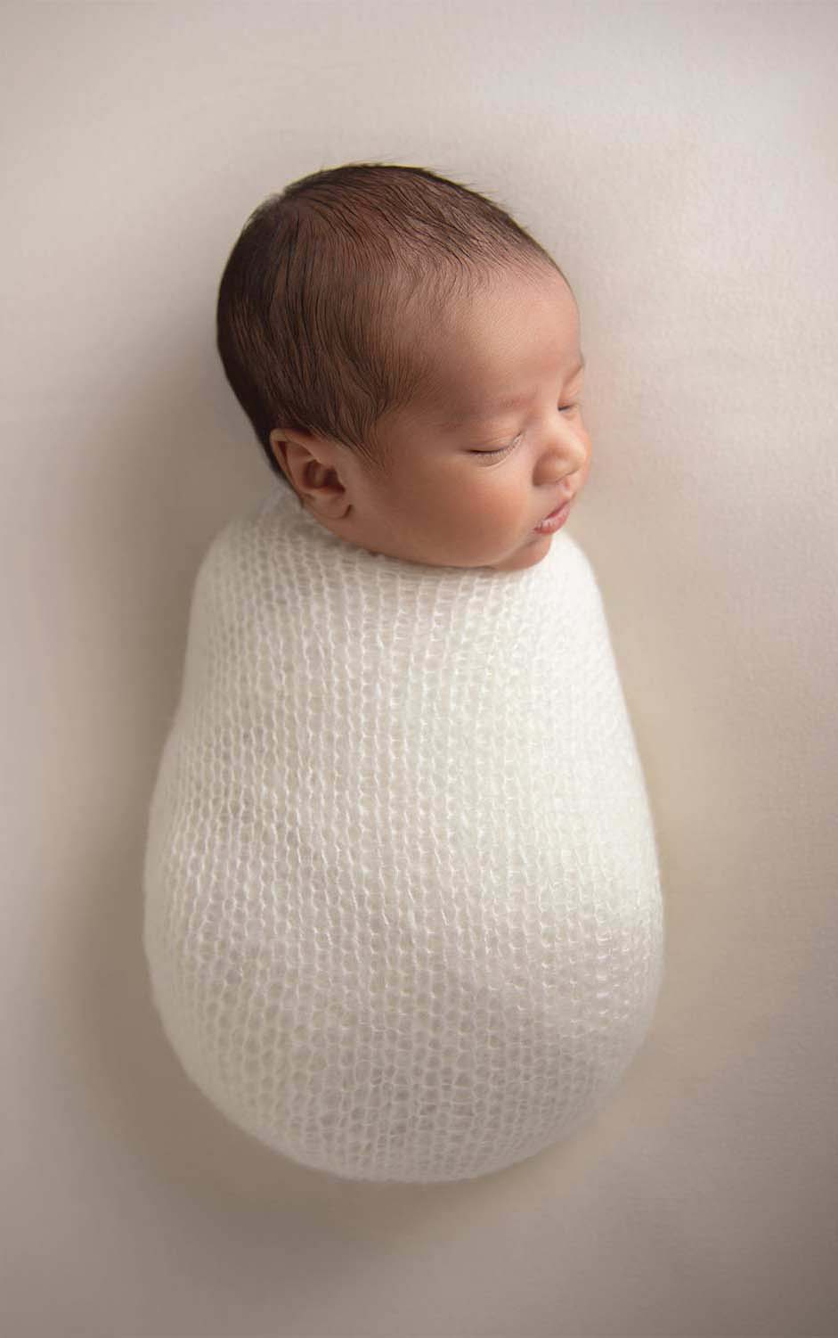 A newborn baby peacefully sleeps wrapped in a soft, white knitted blanket against a light background, captured beautifully by an NYC newborn photographer. The baby's eyes are closed, and their head is gently turned to the side.