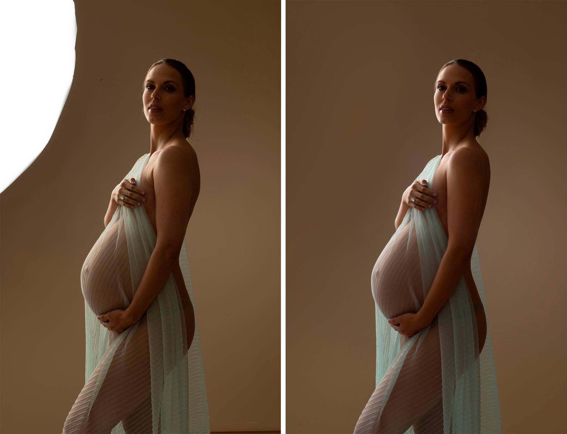 A pregnant woman stands gracefully in a dimly lit studio, her silhouette elegantly captured by a talented maternity photographer. Draped in translucent light blue fabric, she gently holds it over her chest, softly illuminating her baby bump and creating delicate shadows on her figure.