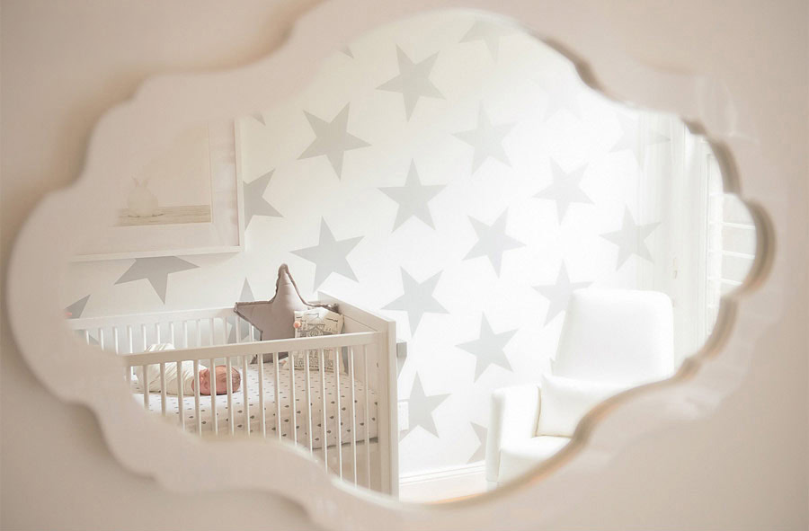A mirror reflects a cozy NYC home nursery with a white crib, star-patterned walls, and a soft armchair. A stuffed toy is placed on the crib, and light filters in through a window, creating a peaceful atmosphere.
