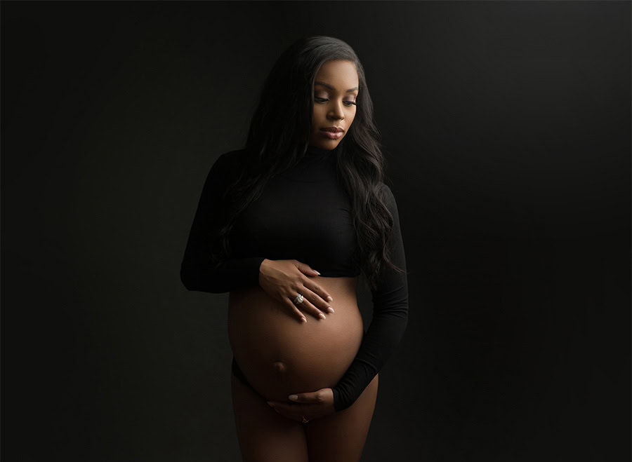 A pregnant woman stands against a dark background, wearing a black long-sleeve top. Captured by a maternity photographer, she gently cradles her bare belly with both hands, looking downwards with a serene expression. Her long hair cascades over her shoulders.