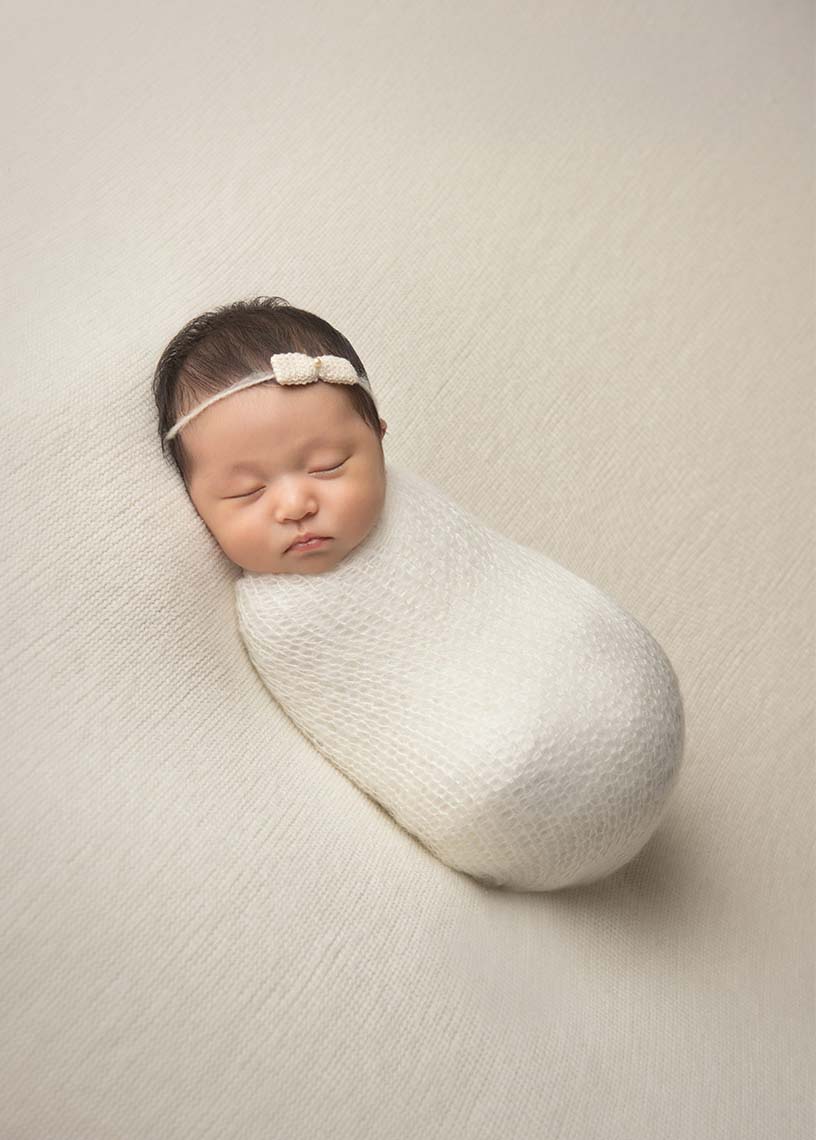A newborn baby sleeps peacefully, wrapped in a soft, creamy-white knitted blanket. Captured by an NYC newborn photographer, the baby wears a delicate headband with a small bow, resting against a matching textured background.