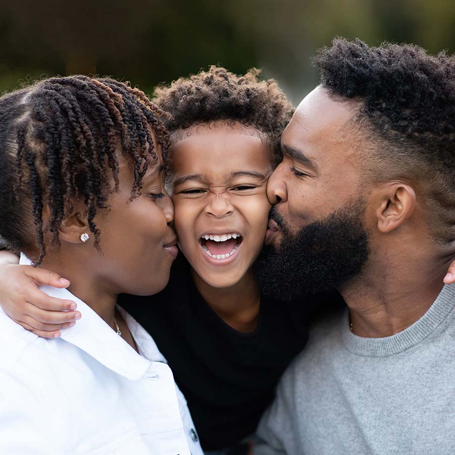 Family photo of a Mom and Dad kissing their son, who is smiling in excitement.