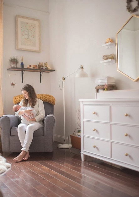 A woman sits in a grey armchair in her modern NYC apartment, cradling a baby. The softly lit room features white walls, wooden floors, and a sleek dresser adorned with decor. A floor lamp and hexagonal mirror complete the calm and cozy atmosphere.