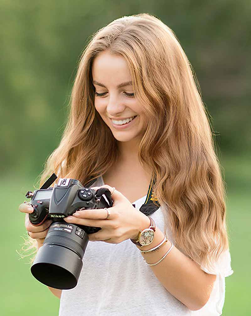 A woman with long, wavy hair smiles while looking at a camera in her hands. She is wearing a white shirt and several bracelets. The background is a blurred green field, suggesting an outdoor setting.