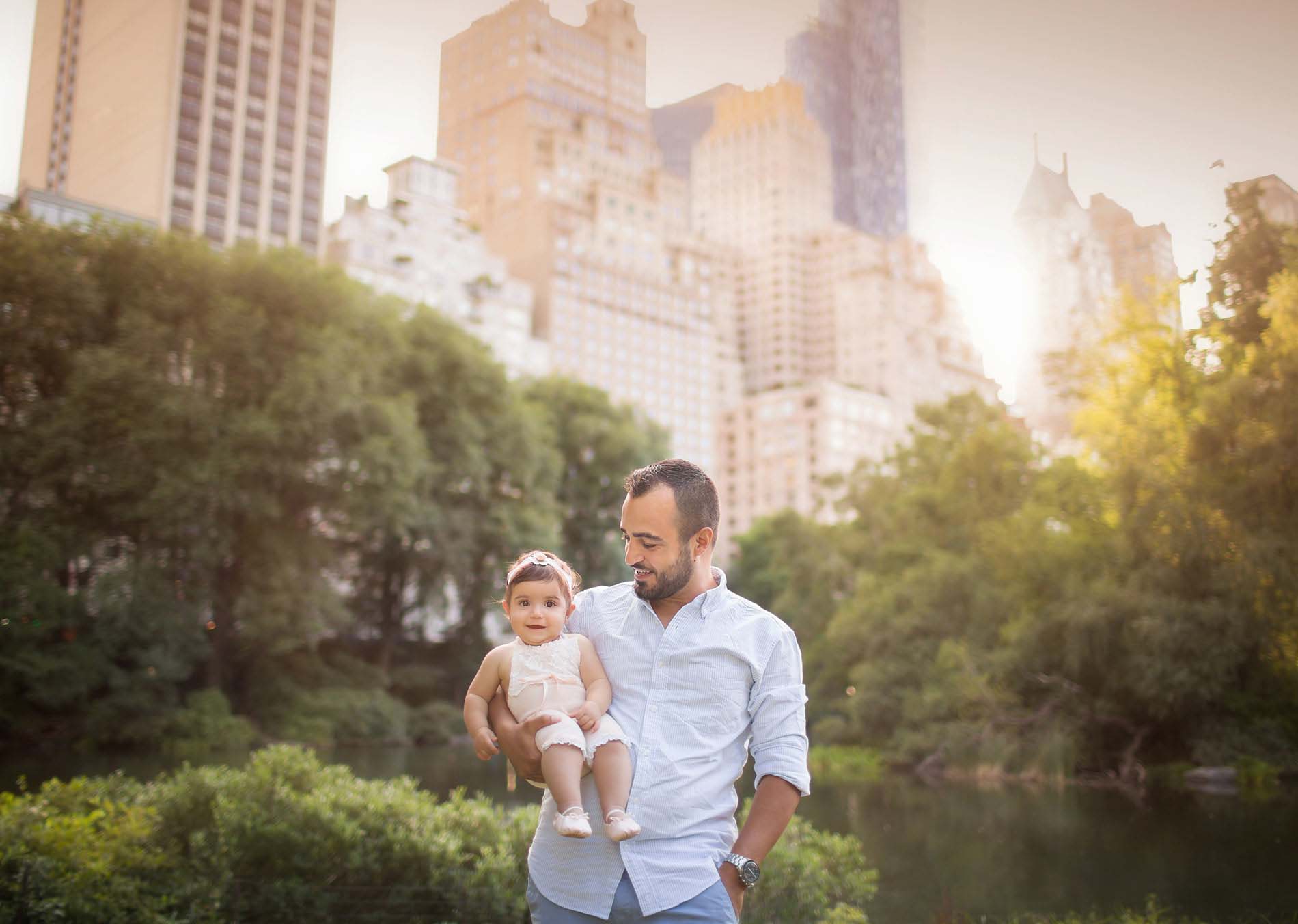 A man in a light blue shirt, captured by a talented Central Park photographer, holds a smiling baby in a headband and light-colored clothes. They stand amidst trees and tall buildings under the sunny sky.
