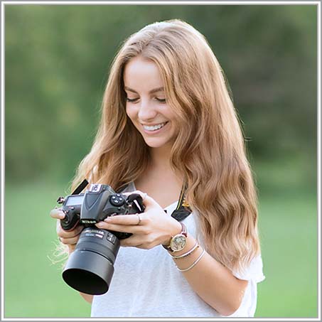 A person with long, wavy hair is smiling while looking at a camera they are holding. They are wearing a white shirt and standing outdoors, with a blurred green background suggesting a natural setting.