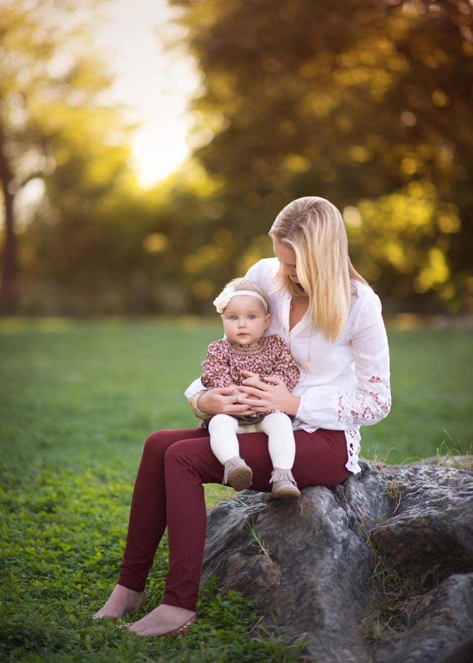 A woman with blonde hair sits on a tree stump in Central Park’s grassy field, holding a baby girl in her lap. The baby wears a floral dress and white tights. The woman looks down at the child, who gazes toward the camera.