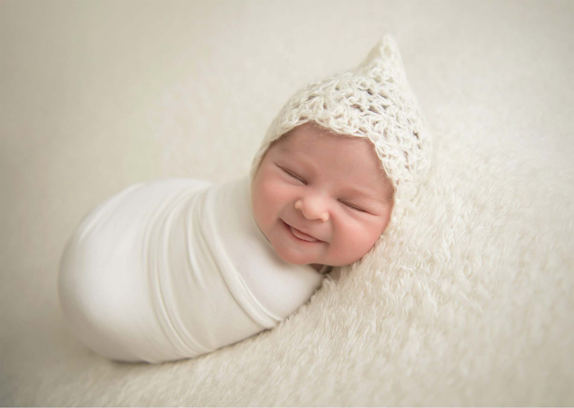 A newborn baby wrapped in a soft white blanket and wearing a knitted white hat smiles while lying on a fluffy white surface.