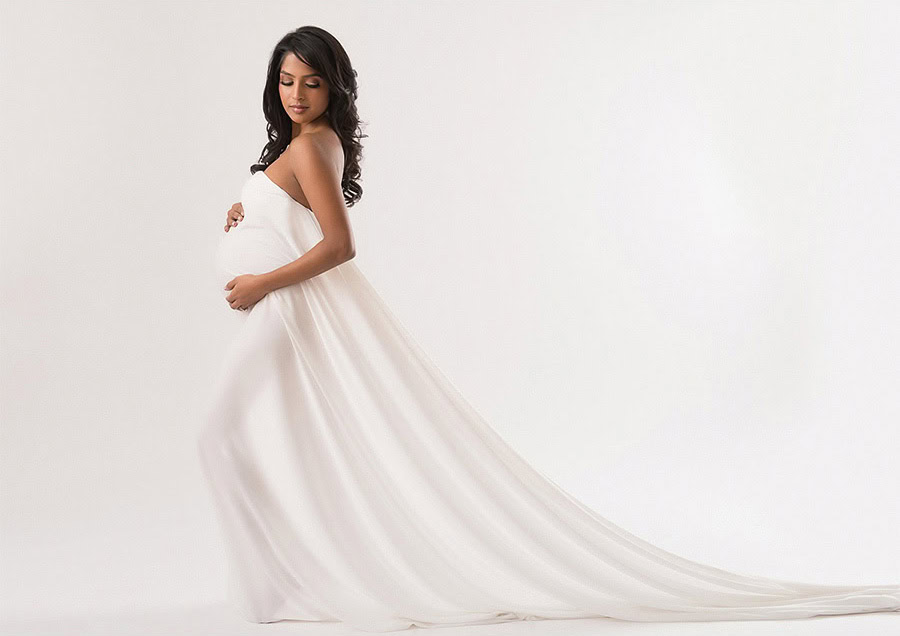 A pregnant woman in a flowing, strapless white gown stands against a plain white background at an NYC photography studio, gently cradling her belly and looking down with a soft smile.
