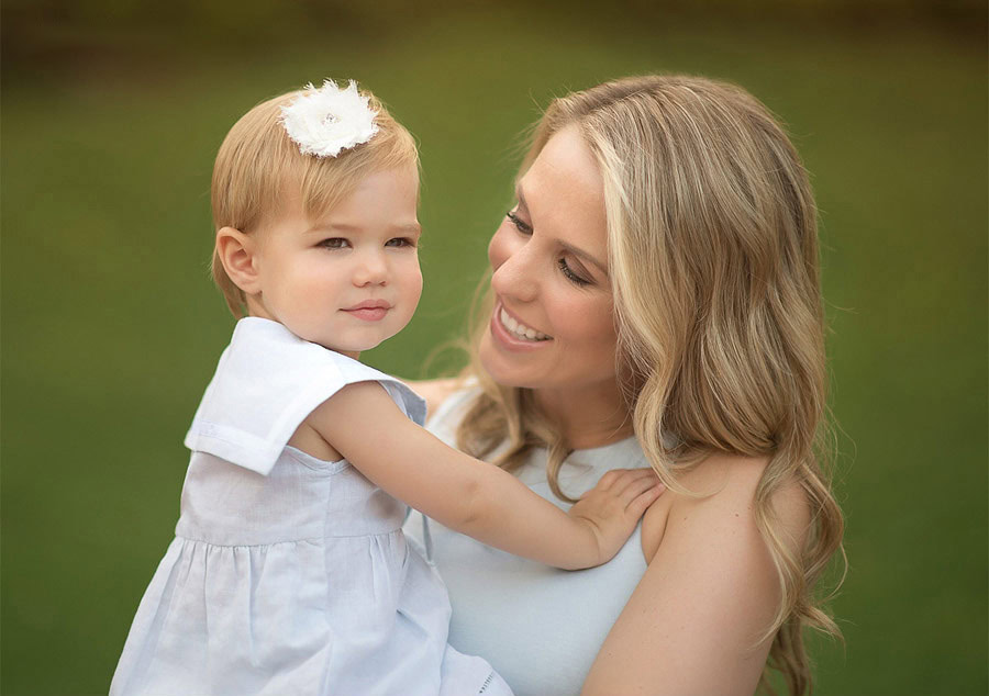 A woman with long blonde hair smiles while holding a young girl dressed in white with a white flower headband, standing outdoors against a blurred green background.