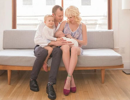 A modern NYC family of four sits on a light gray sofa. The father and mother, both with light hair, cradle a newborn as an older child rests on the father's lap. They all share a tender moment together.