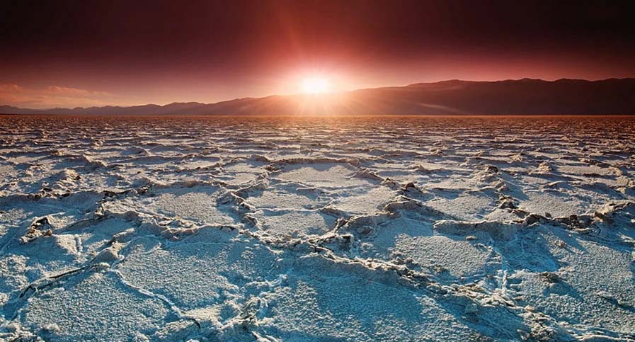 The sun sets over distant mountains, casting a warm glow on the cracked white salt flat of Badwater Basin in Death Valley National Park. The sky transitions from deep blue to orange near the horizon, highlighting this dramatic landscape sunset.