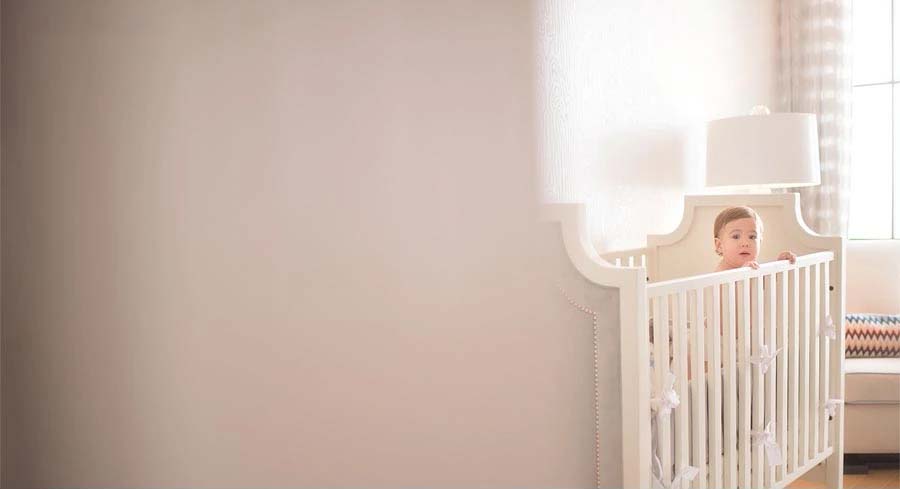 A baby stands inside a white crib near a window in a softly lit room, holding onto the crib railing and gazing outward—a serene moment in a modern NYC apartment with a neutral-colored wall on the left side.