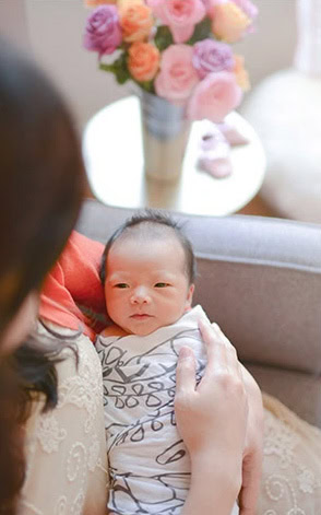 A mother holding her infant, wrapped in a patterned blanket, is cradling the newborn in her arms. In the softly blurred background, a vase of colorful flowers rests on a table.