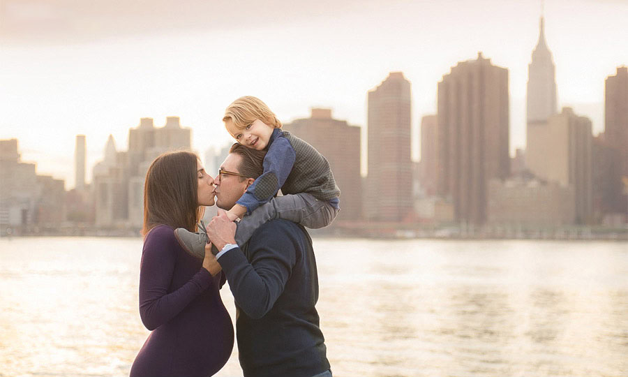 A pregnant woman and a man kiss while the man carries a smiling child on his shoulders. The family stands by the water with a city skyline, including skyscrapers and the Empire State Building, in the background at sunset.