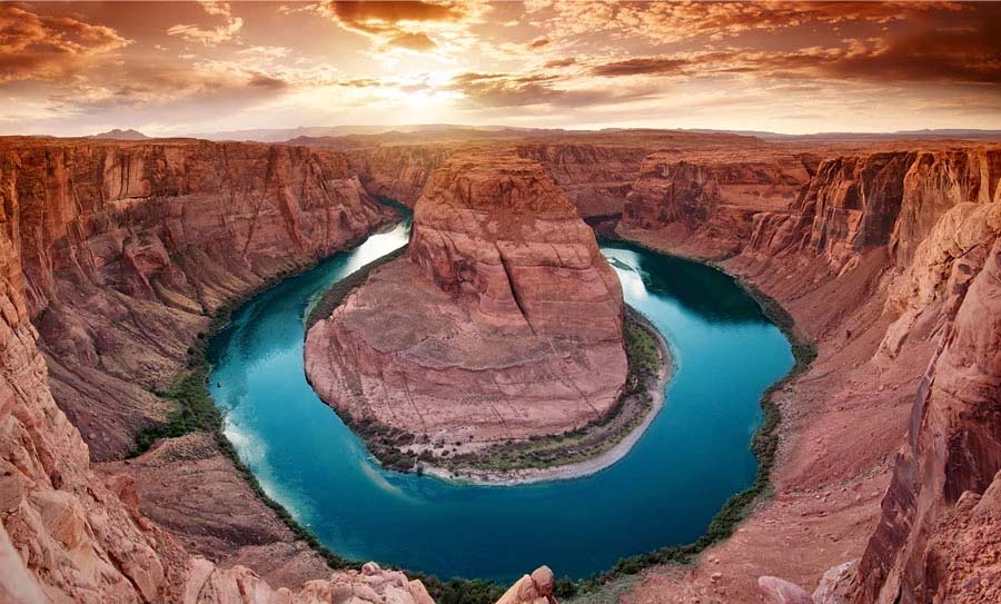 A wide view of Horseshoe Bend shows a river curving around a large rock formation, surrounded by steep, rocky canyon walls at sunset under a partly cloudy sky, highlighting the dramatic beauty of this iconic landmark.