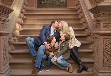 A couple sits on outdoor brownstone steps in the Upper West Side, kissing, while two young boys sit in front of them, looking up and smiling. The family appears happy and relaxed in a casual setting.