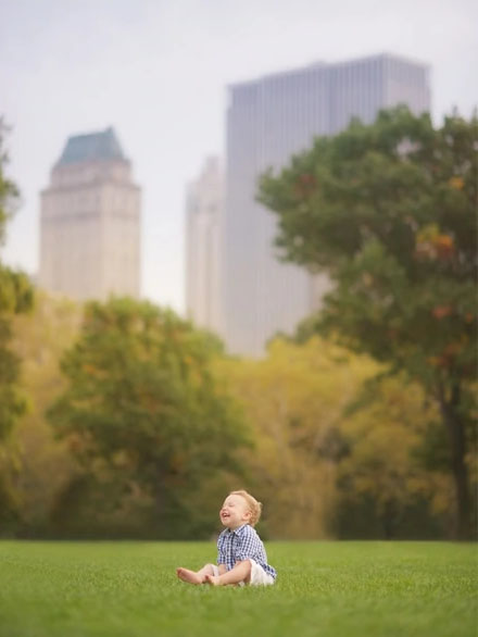 A small child sits barefoot on green grass in a park, smiling and looking up. Captured by a talented baby photographer, tall buildings and trees blur in the background, hinting at an urban setting.