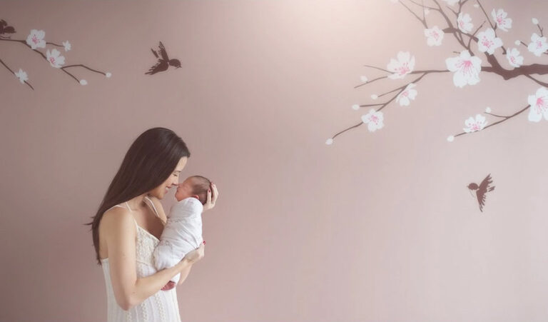 A woman in a white dress gently holds a newborn baby wrapped in a blanket, standing beside a pastel wall decorated with painted cherry blossoms and birds. The scene feels calm and tender.