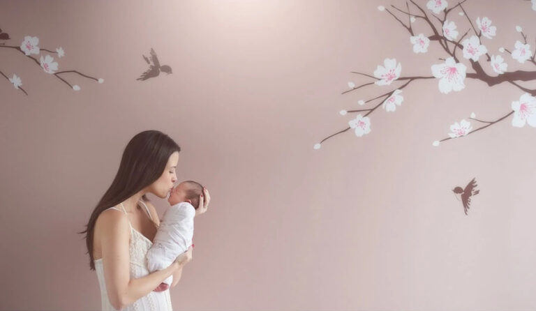 A woman in a white dress gently kisses a newborn wrapped in a blanket, standing near a wall decorated with painted cherry blossoms and birds. The background is soft pink, creating a serene and tender atmosphere.