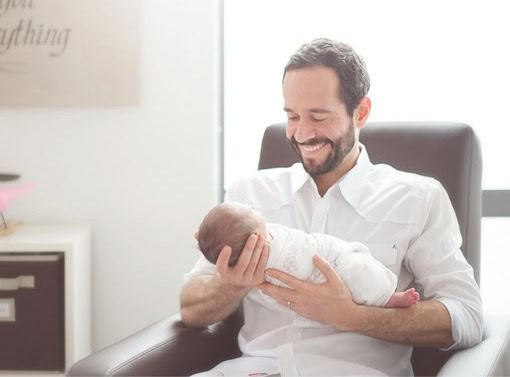 A smiling father with a beard, dressed in a white shirt, sits in a chair and gently cradles his infant son in his arms. The background is softly lit, giving a warm and peaceful atmosphere.